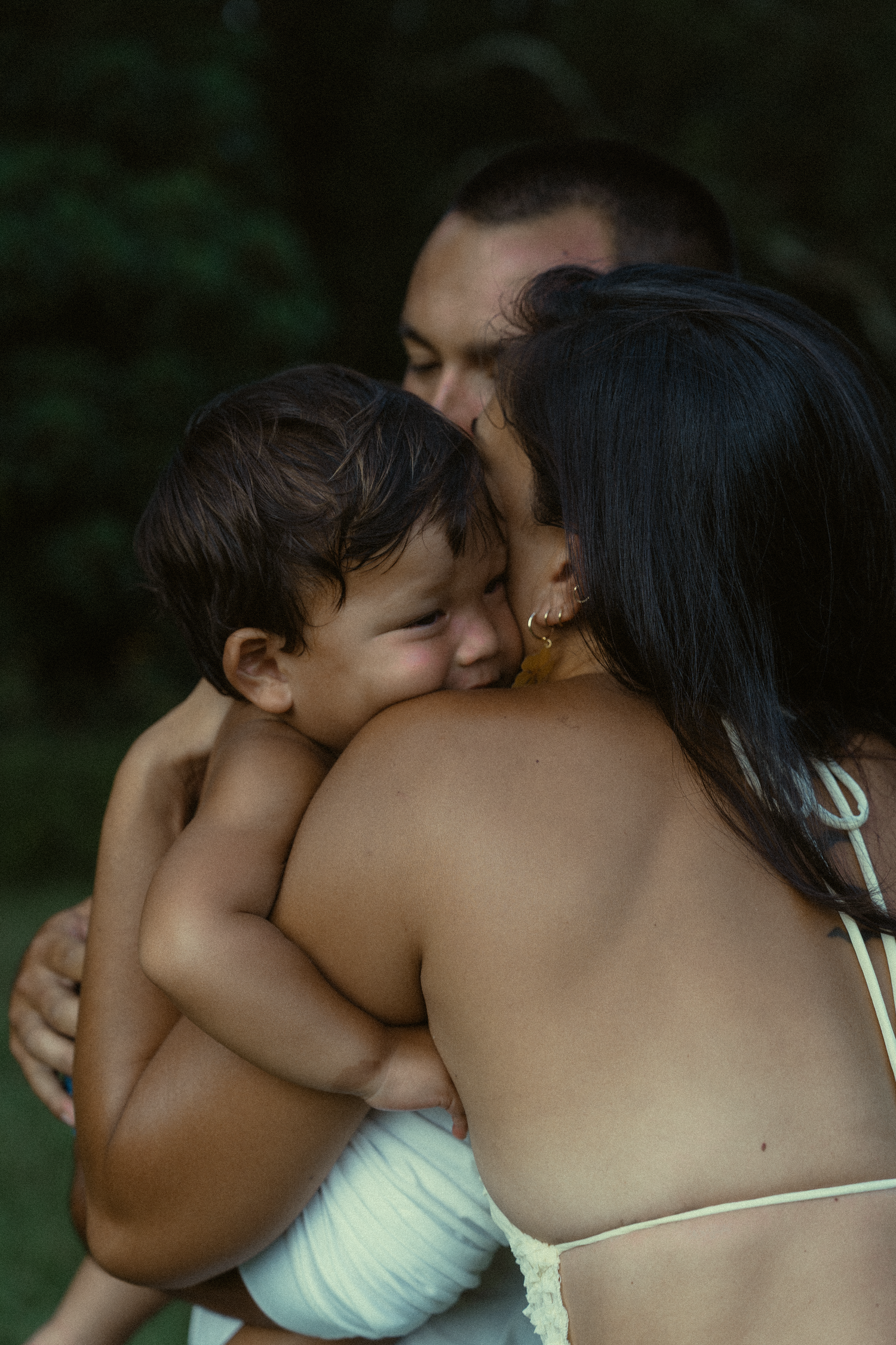 A tender family moment captured outdoors as they share an intimate embrace against a dark green background.