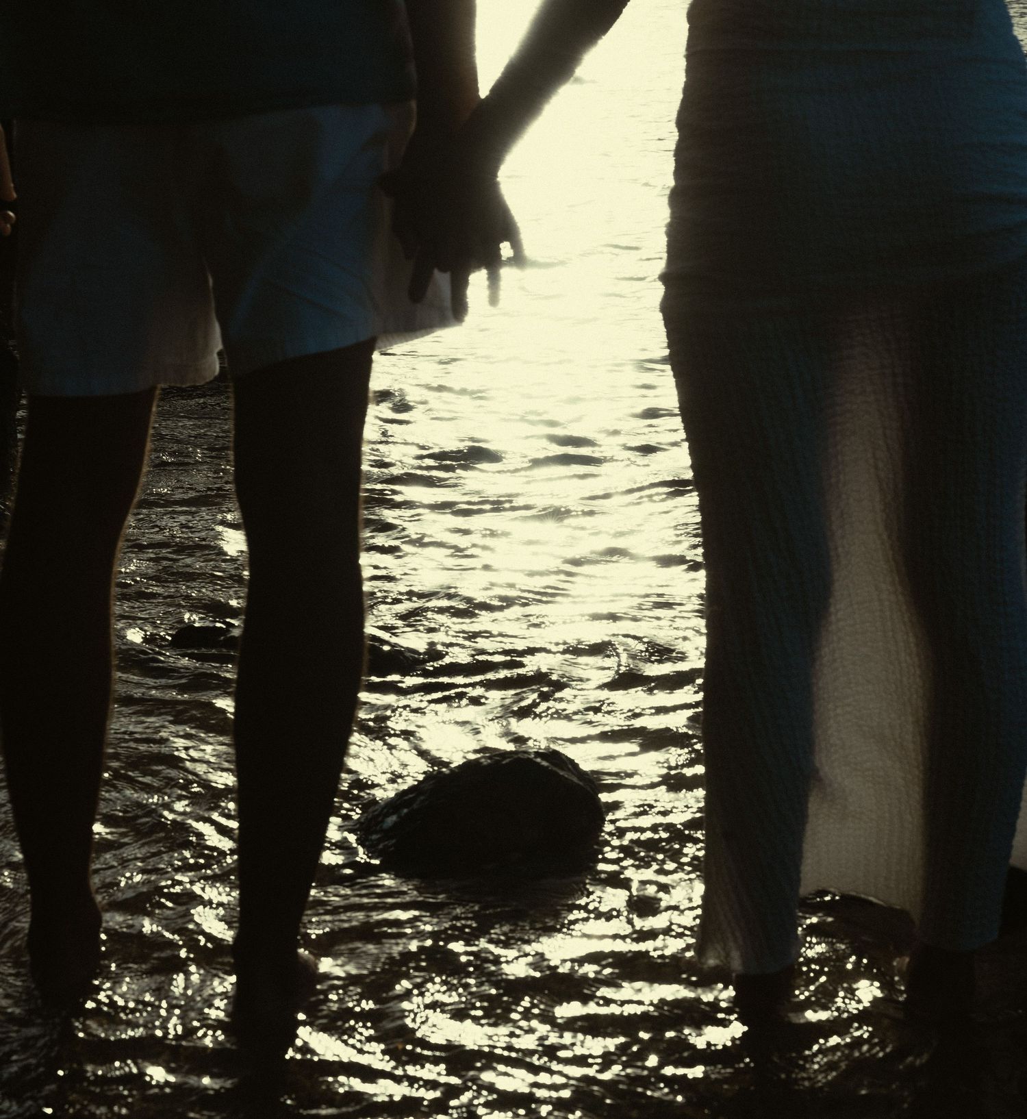 Silhouetted legs of people standing in shallow water at sunset with light reflecting off the rippling surface.