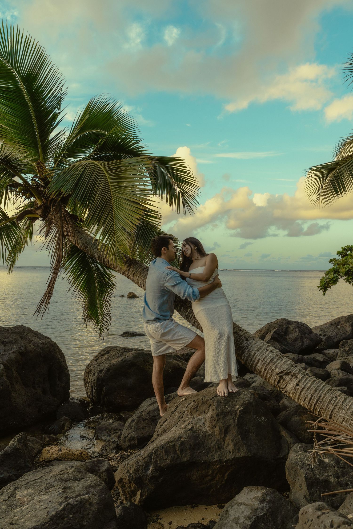 A romantic sunset scene on a tropical beach with palm trees leaning over volcanic rocks by the ocean.