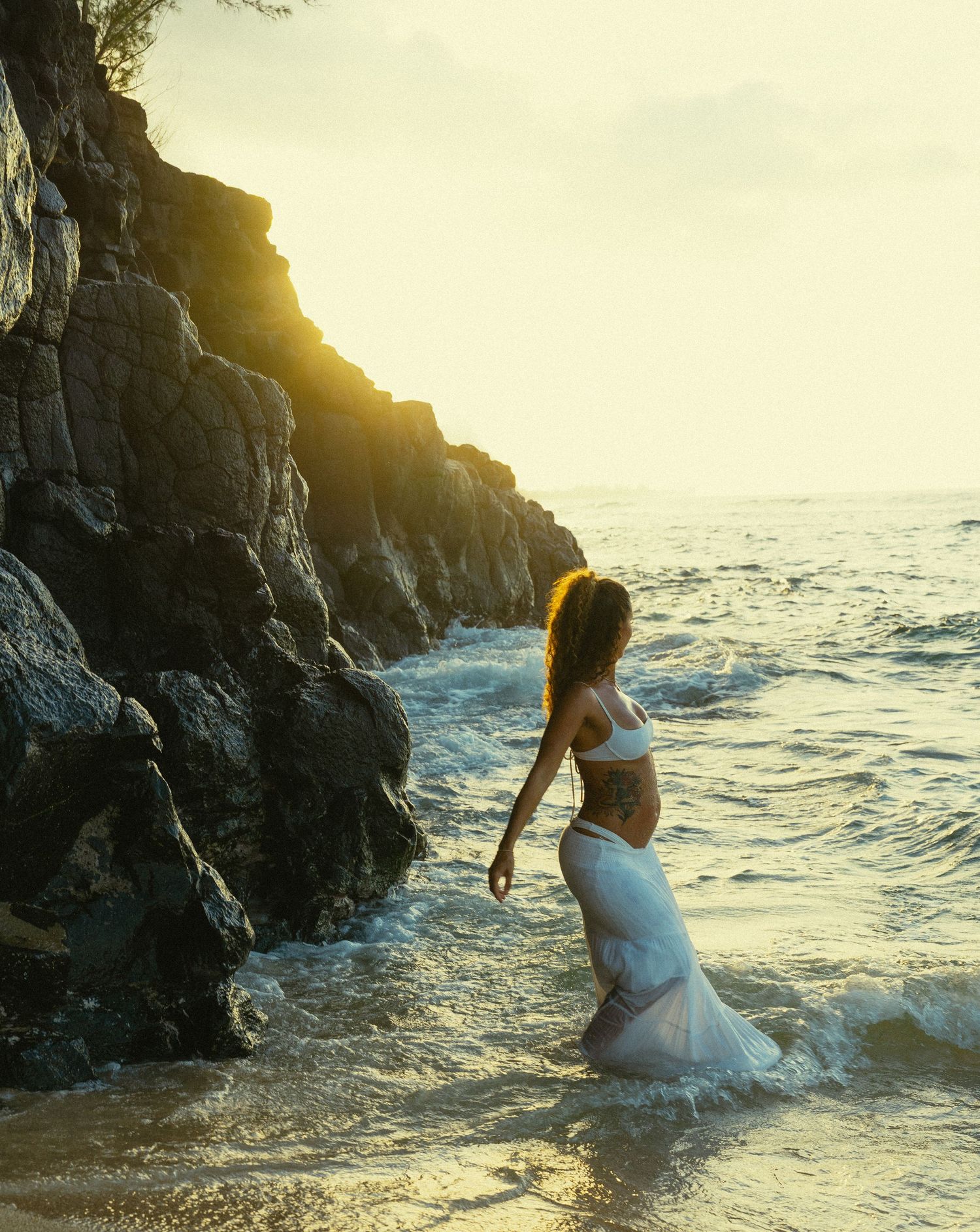 Pregnant woman in white dress sitting by ocean rocks at sunset with waves crashing behind her.
