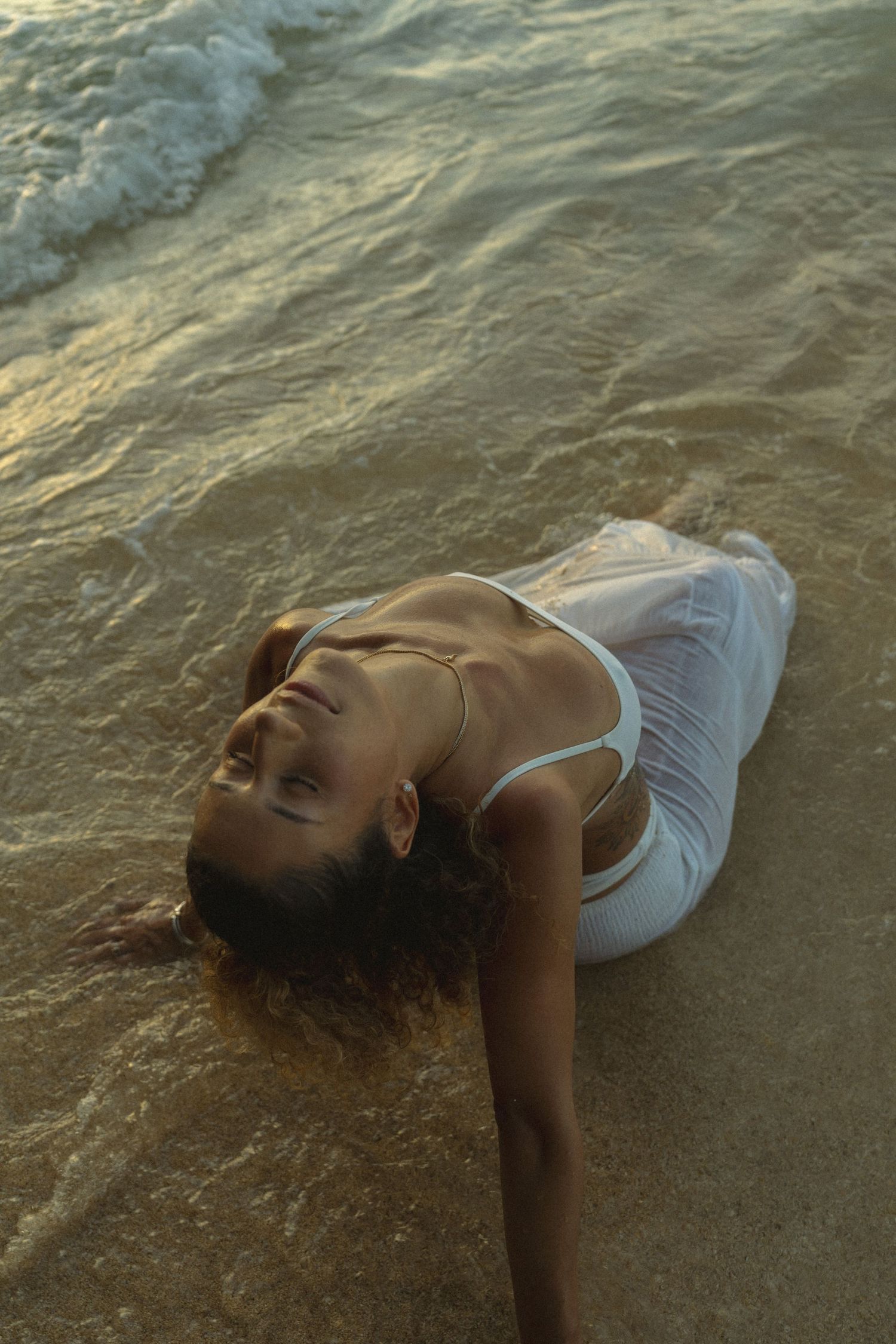 Figure in white clothing lying artistically on wet sand at the beach during golden hour.