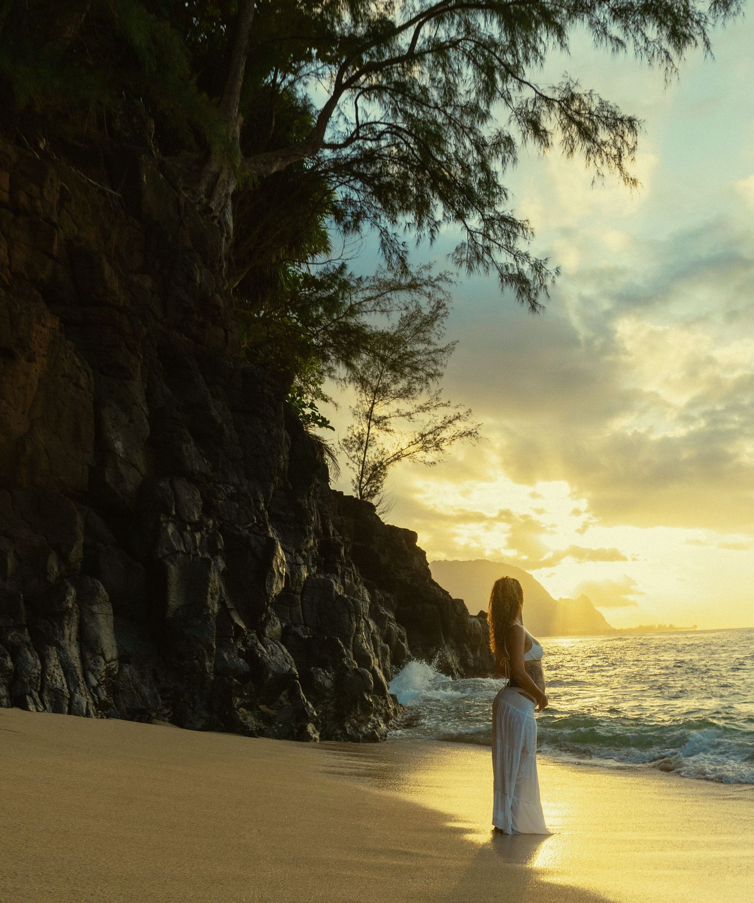 A figure in white stands on a sandy beach at sunset with dramatic cliffs and ocean waves in the background.