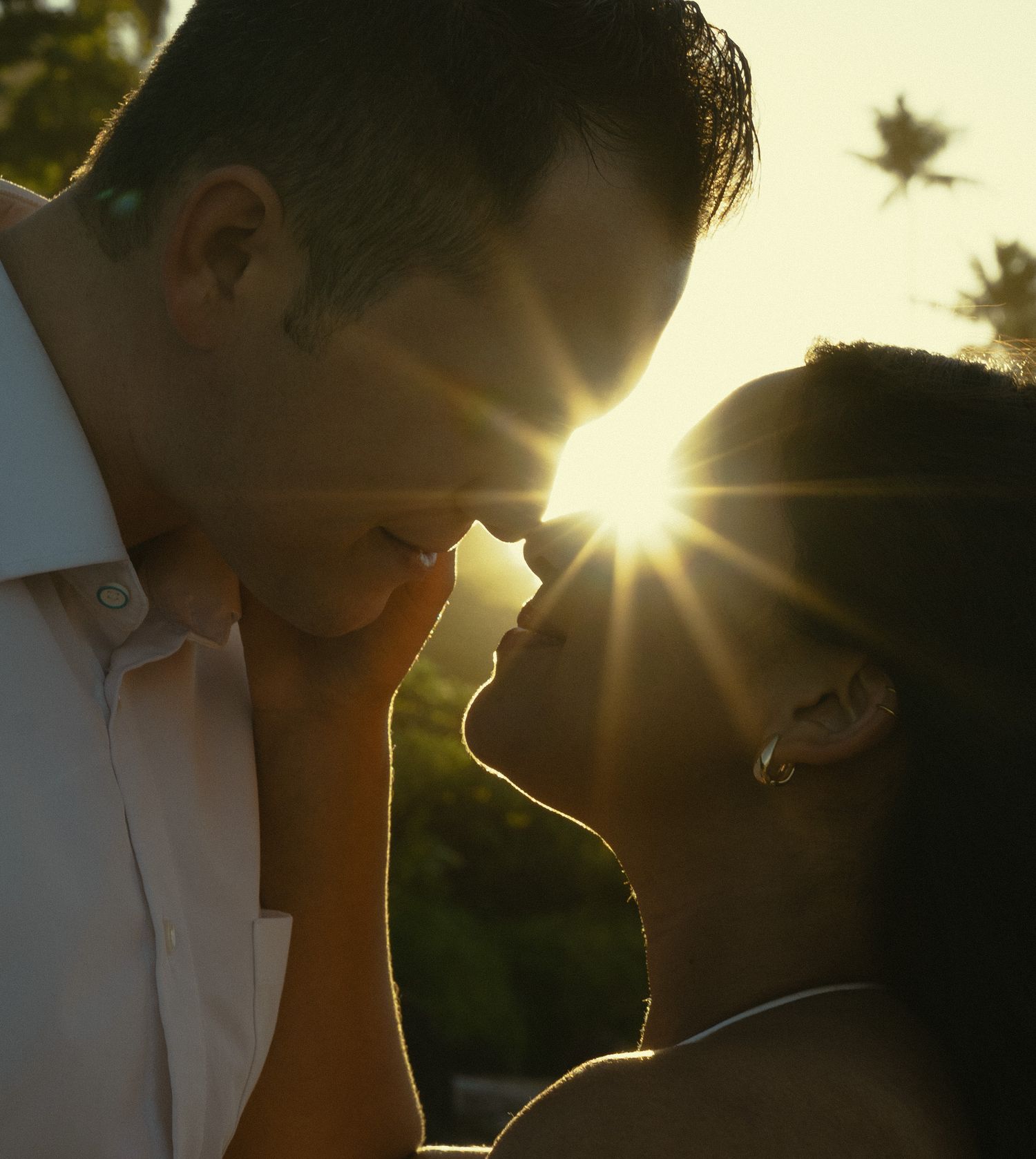 Silhouetted couple share an intimate moment at sunset with palm trees and sunburst in the background.