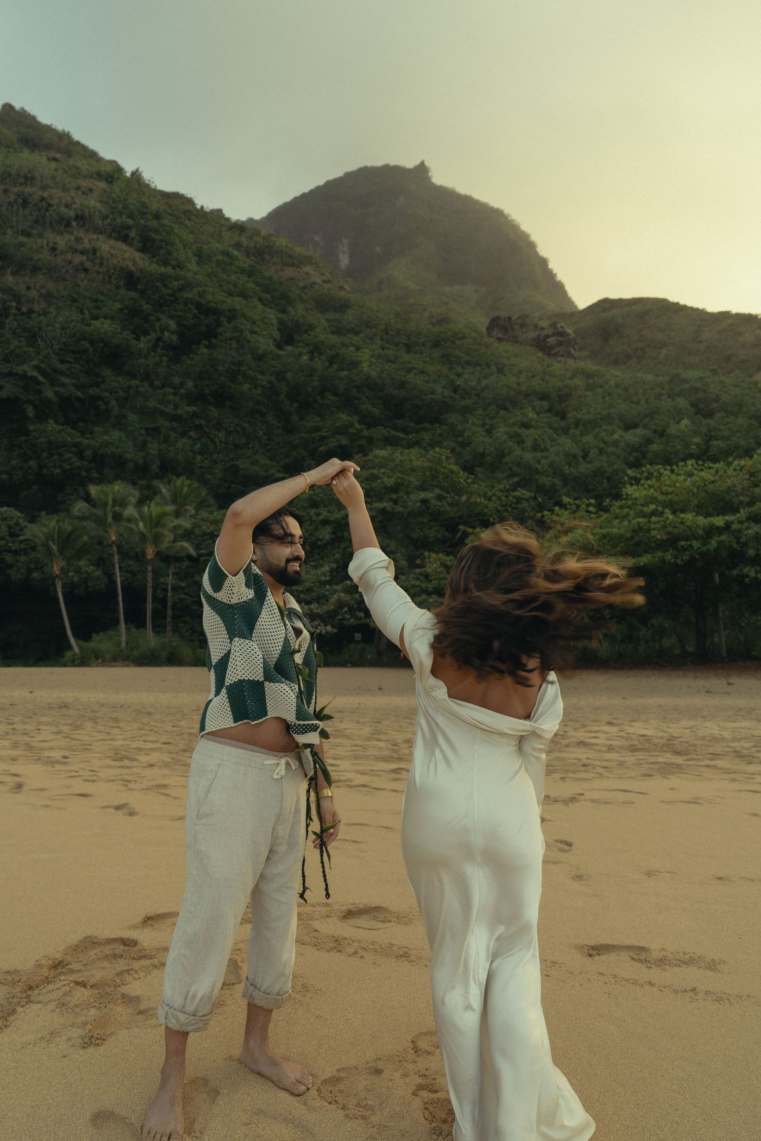 A couple in white outfits dances together on a scenic beach at sunset with mountains in the background.