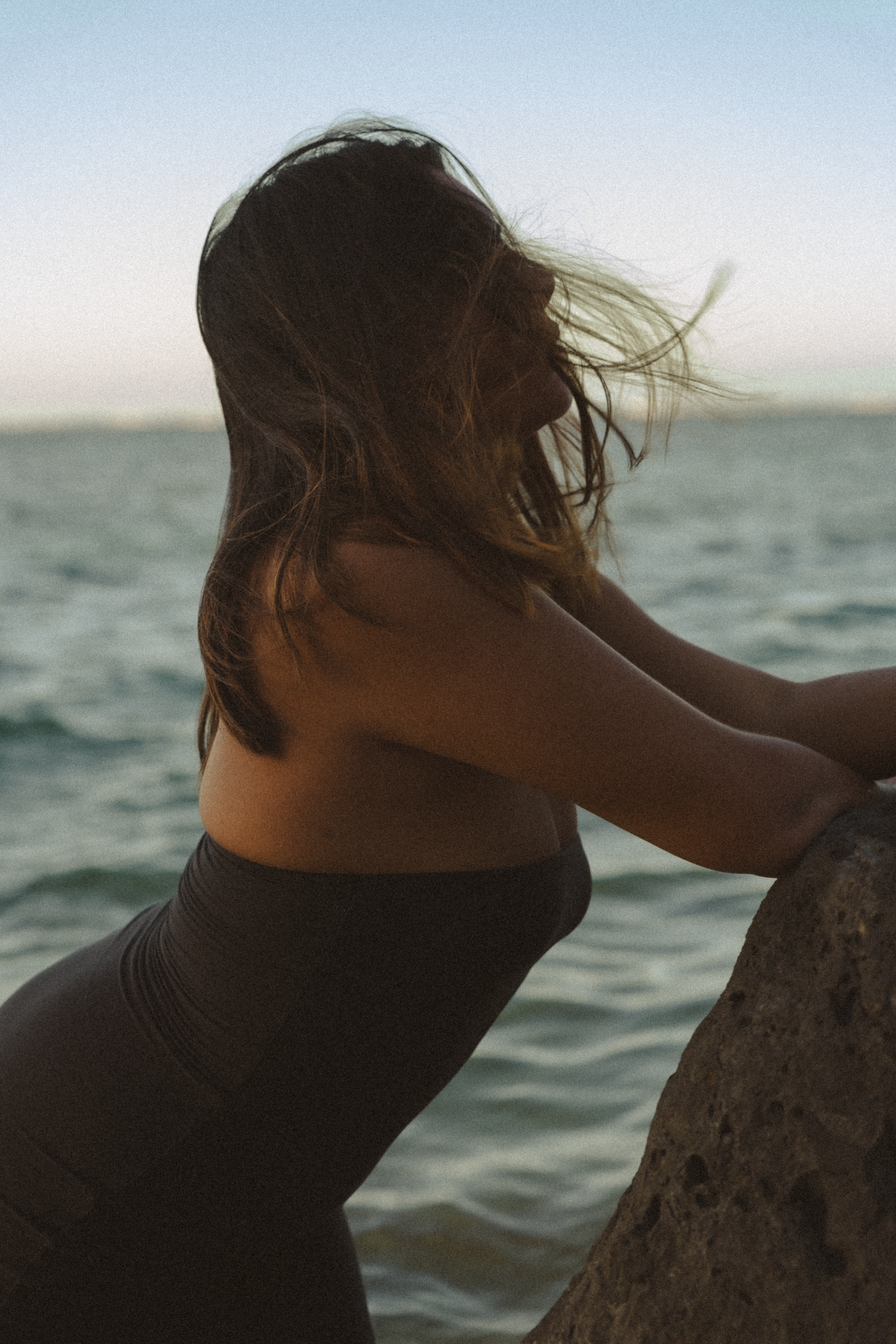 Silhouetted figure against ocean waves at sunset with wind-blown hair.
