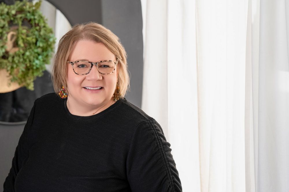 Professional headshot of someone wearing glasses and a black sweater smiling warmly against a light background with greenery.