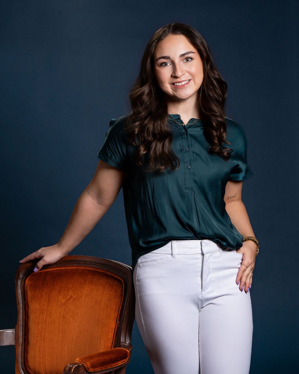 Professional studio portrait against dark blue backdrop with wooden chair and casual business attire.