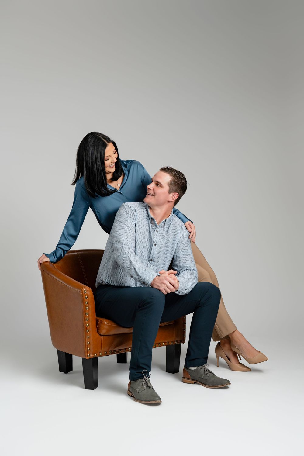 A couple shares an intimate moment while sitting together in a brown leather armchair against a gray backdrop.
