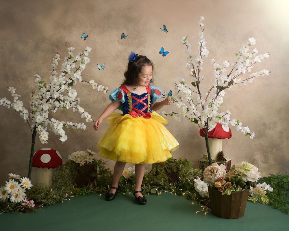 Child in Snow White costume poses in whimsical garden setting with mushrooms and butterflies.