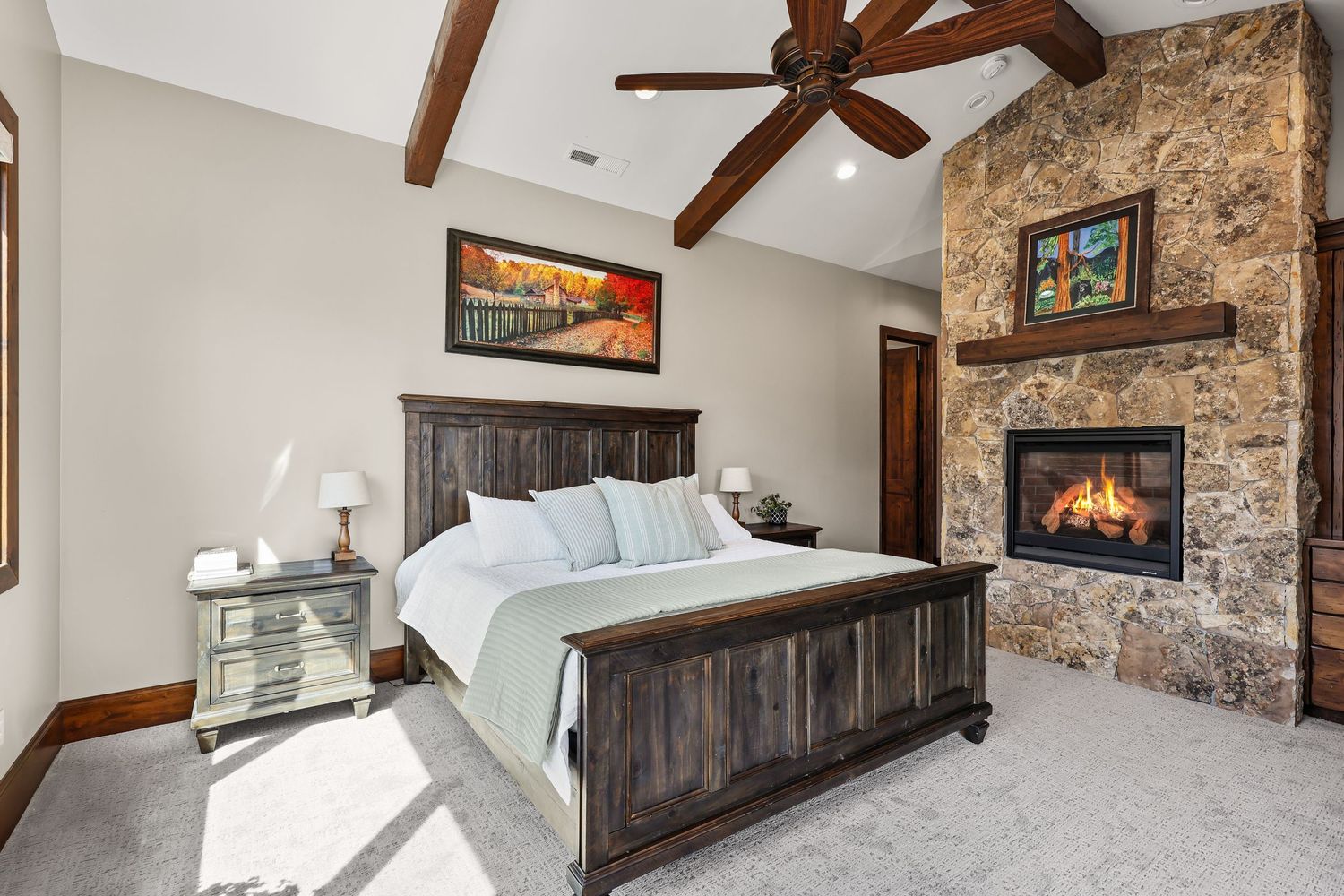 Rustic bedroom with stone fireplace, dark wood bed frame, vaulted beam ceiling, and carpet floors. Captured by Slate Media (best in UT)