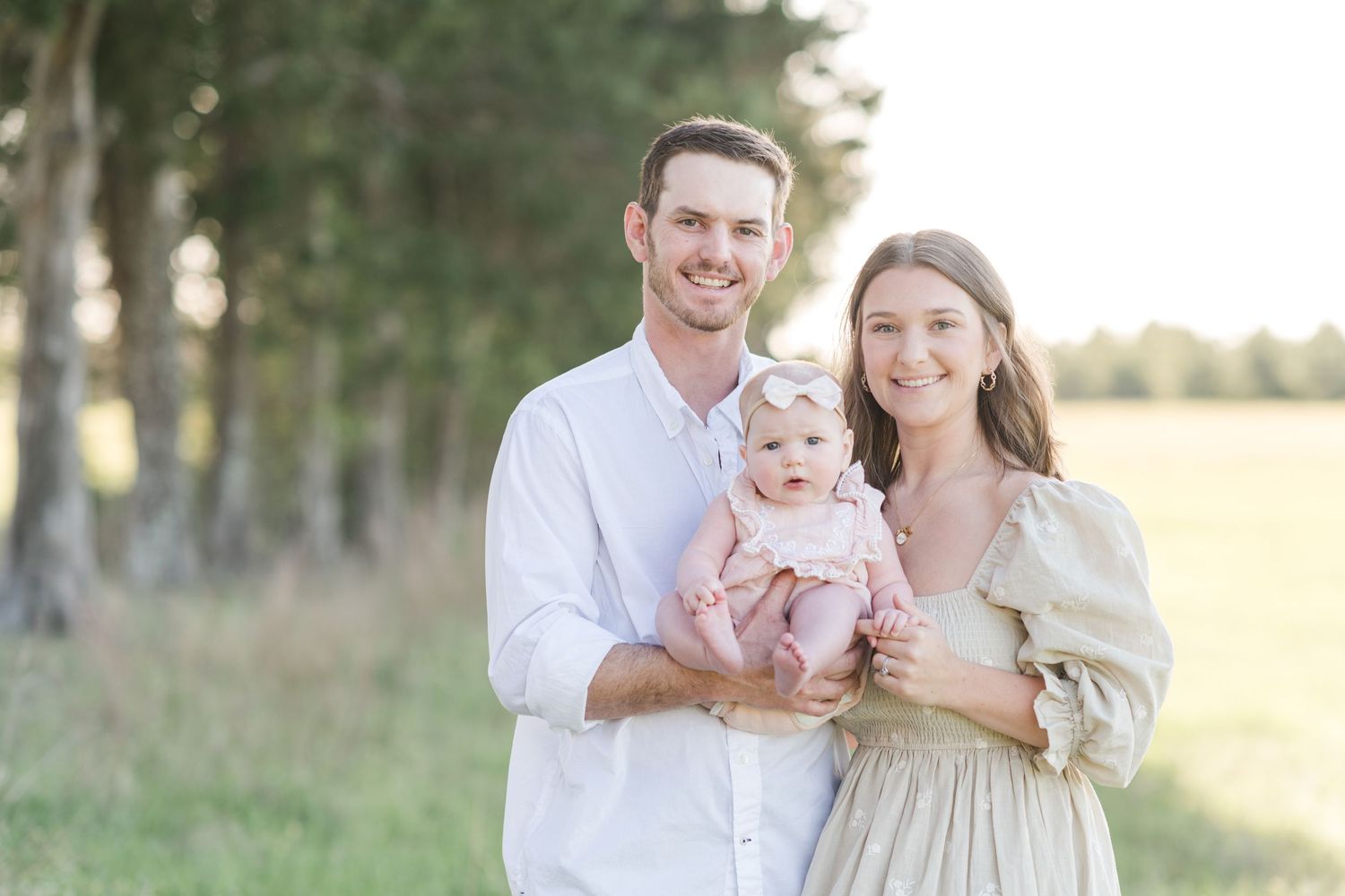 A smiling young couple holds their baby girl in a pink outfit in a sunlit open field.