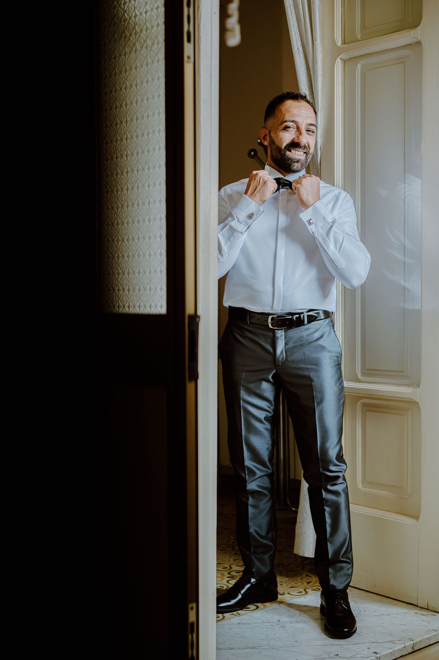 A person in a white shirt and gray pants adjusts their bowtie in a doorway.