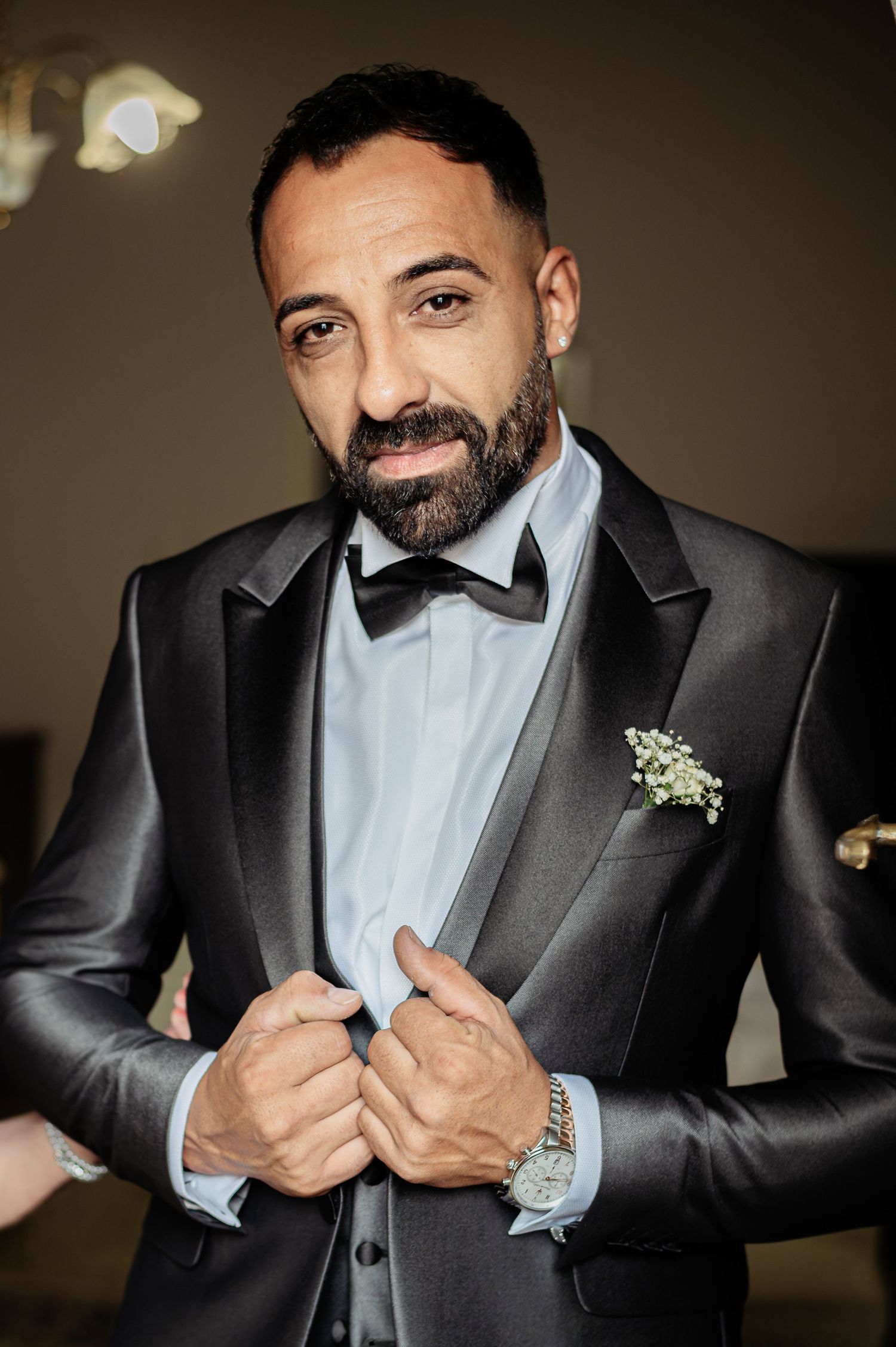 A formal portrait of a person in a black tuxedo with bowtie and boutonniere.