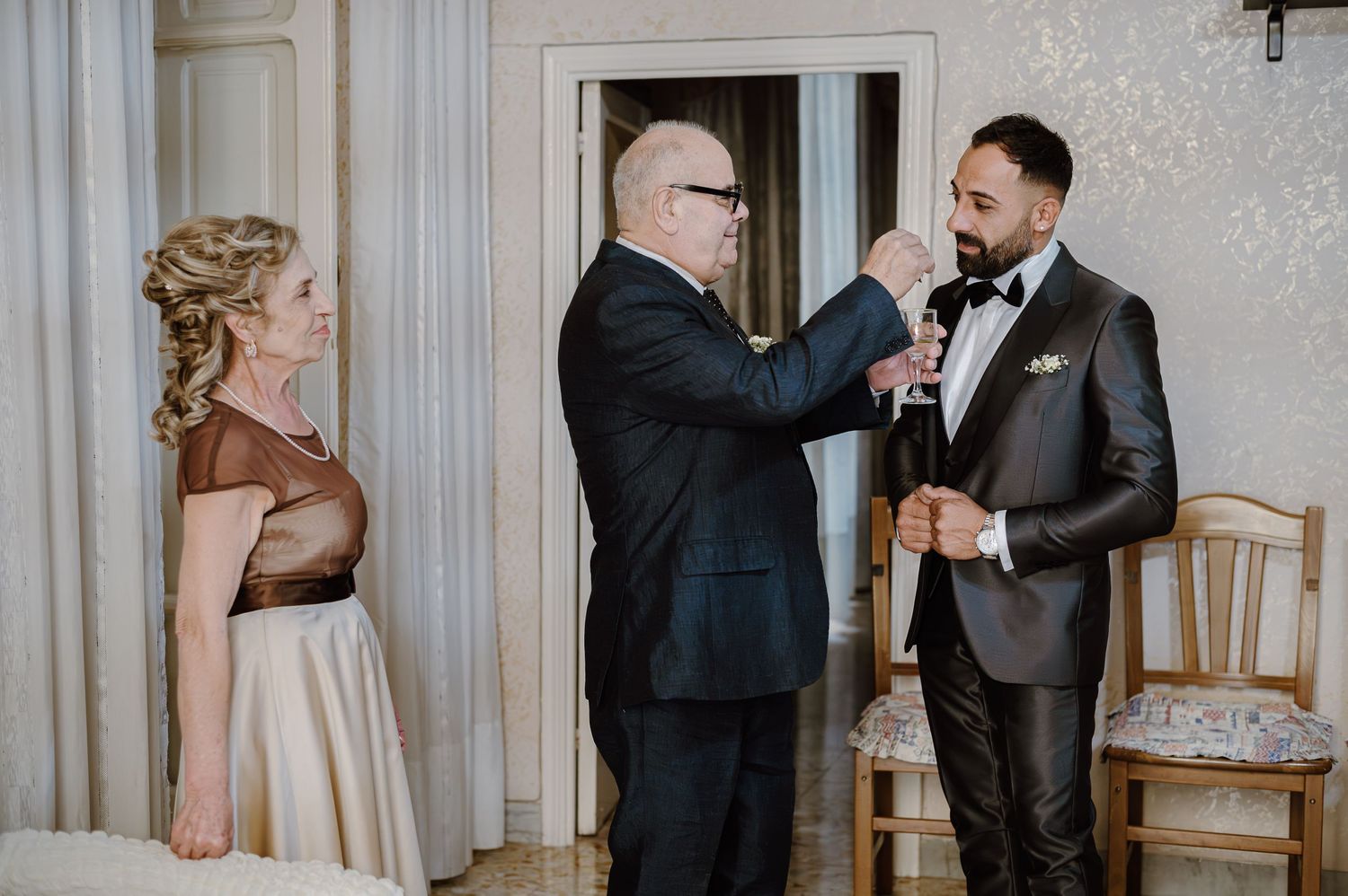 Three people in formal wear interact during a wedding preparation moment.