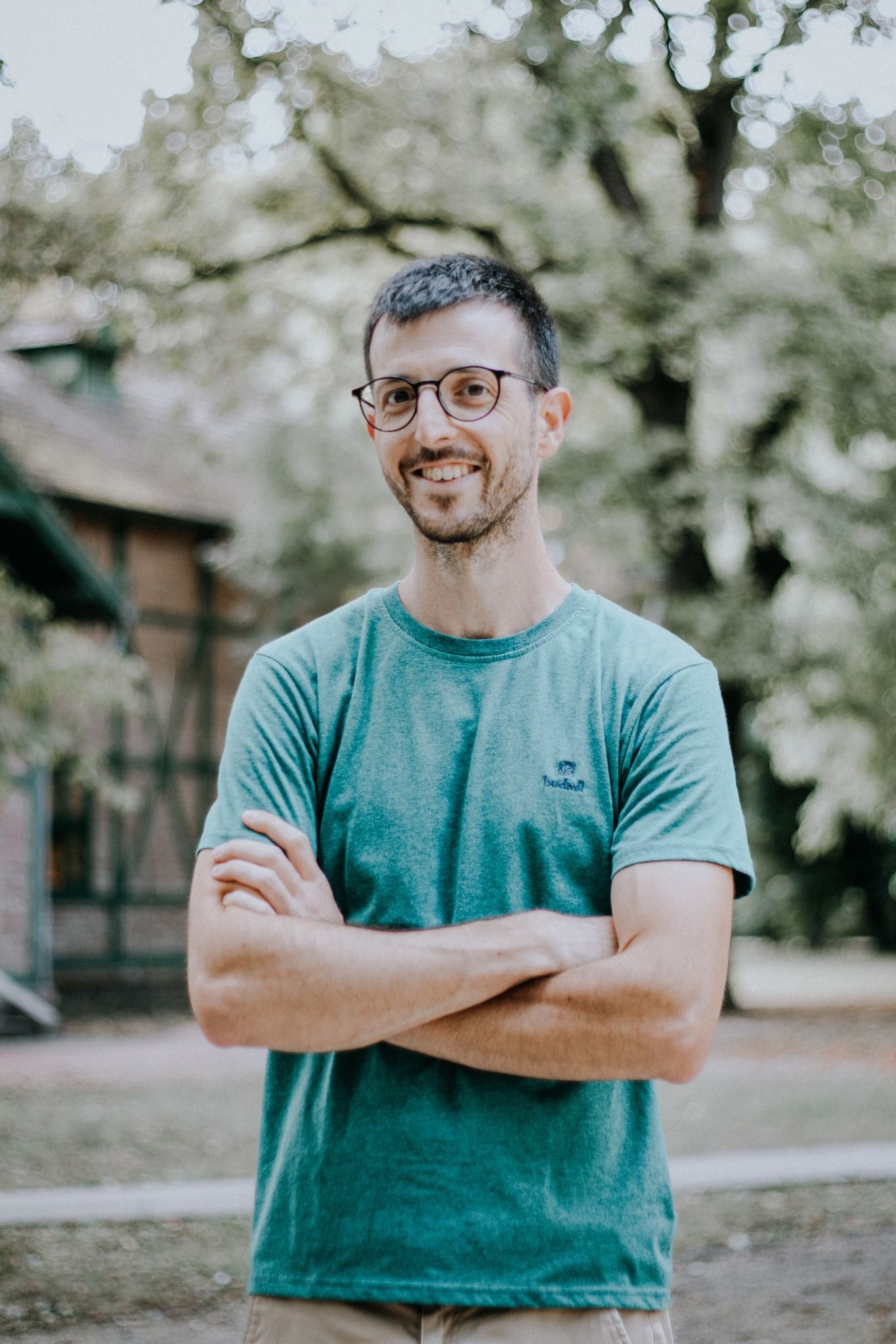 Smiling man with glasses and crossed arms wearing a teal t-shirt outdoors in a park.