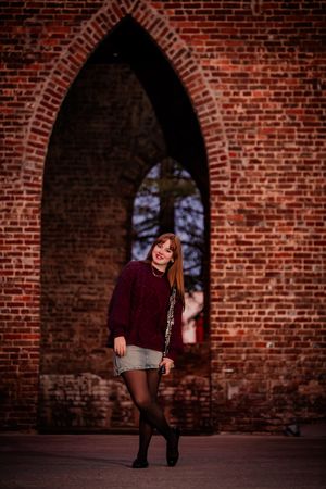 Person in burgundy sweater poses against rustic brick wall with arched window in moody lighting.