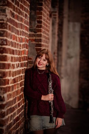 A musician in a burgundy sweater holds a clarinet while standing against a rustic brick wall in moody lighting.