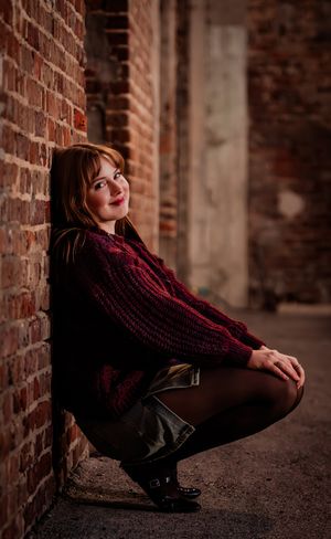 A person in a burgundy sweater poses against a brick wall in a moody urban setting.