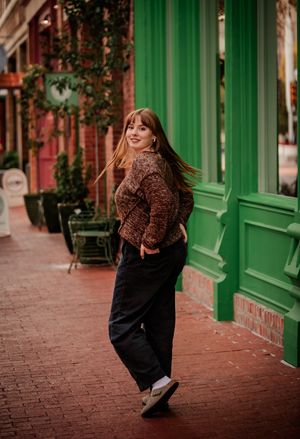 A person in a brown sweater and black pants walks along a brick sidewalk past storefronts with green trim.