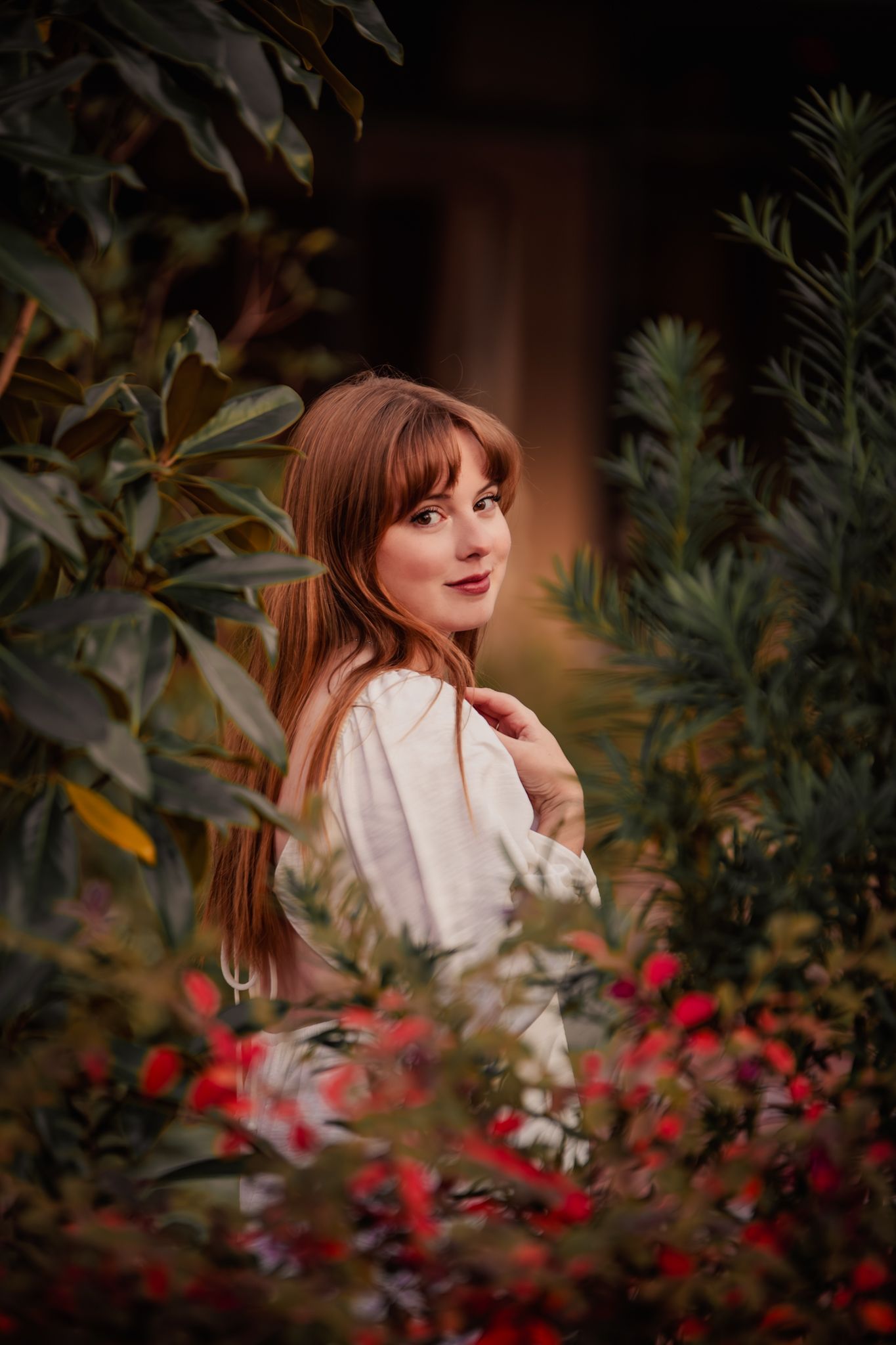 A person in a white top poses among red flowers and green foliage in a garden setting during golden hour.
