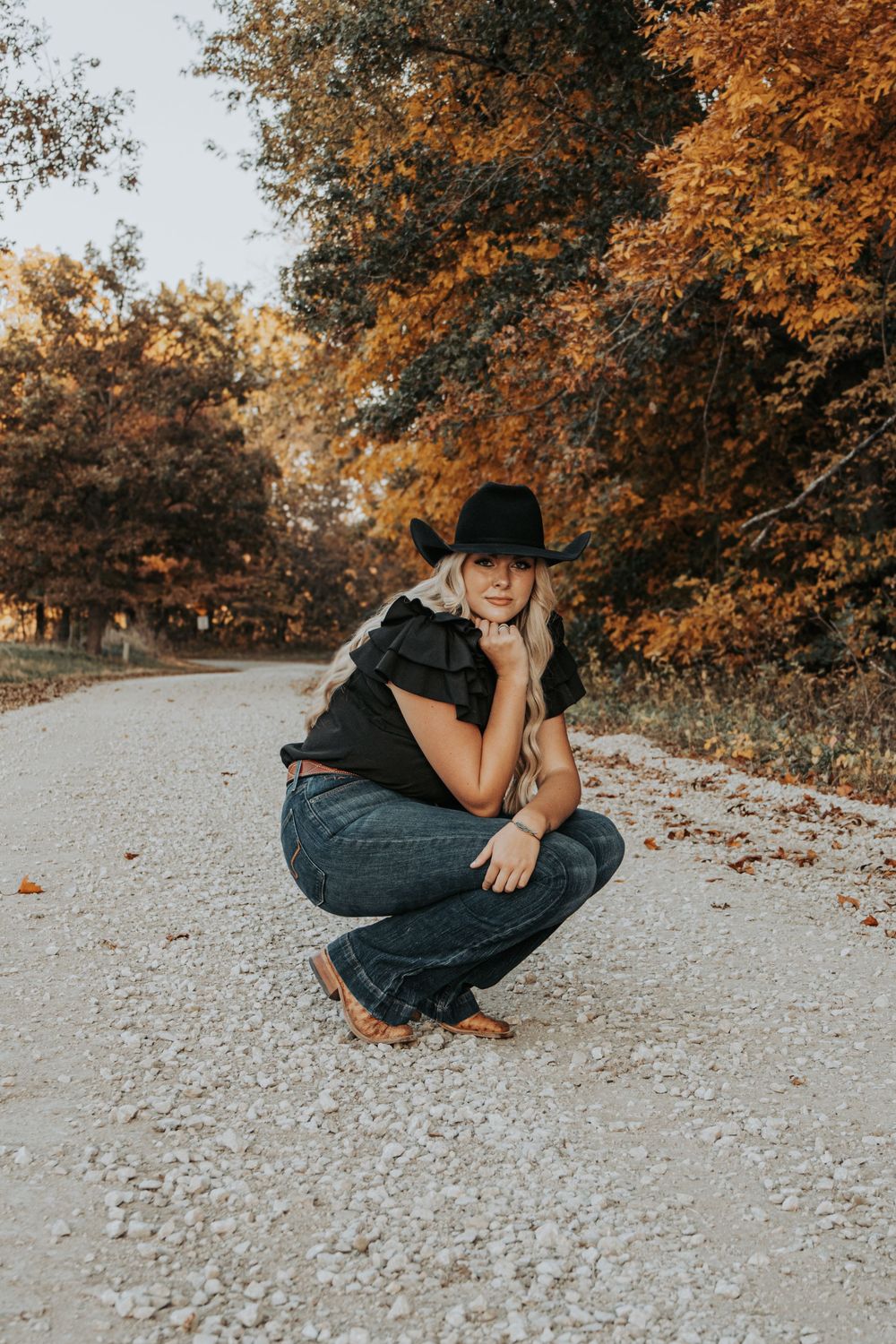 A crouching figure in denim on a rural gravel path with fall foliage in background.