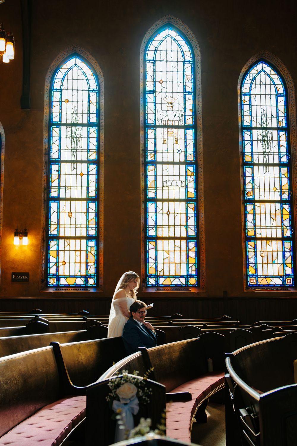 A row of stained glass church windows illuminates wooden pews while a figure lounges dramatically across the seats.