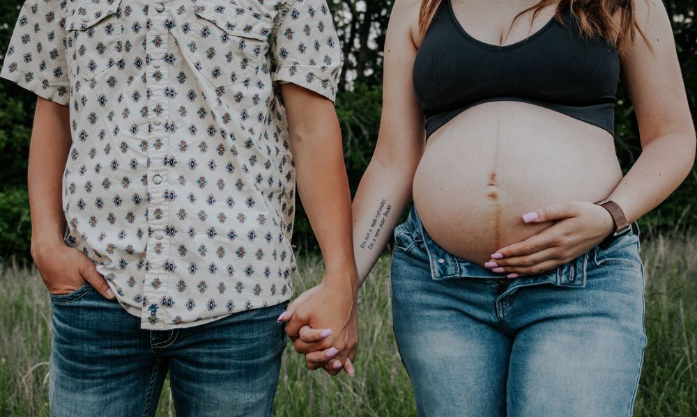 Close-up view of couple holding hands with pregnant belly in jeans and polka dot shirt against outdoor setting.