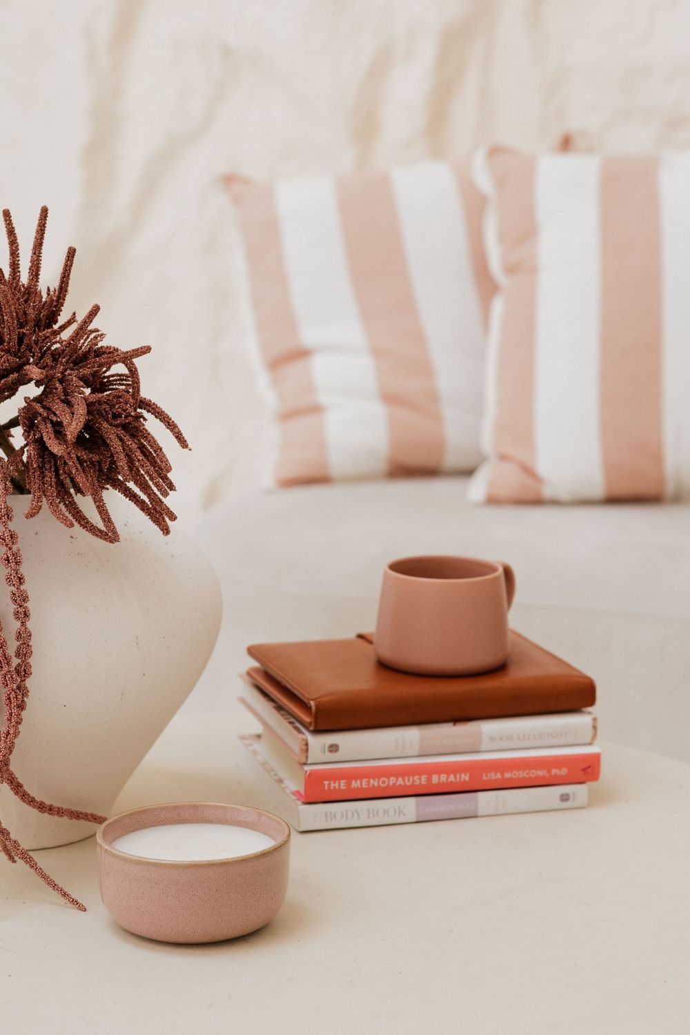 Cozy styled vignette with stacked books including The Menopause Brain, terracotta mug, candle, and white vase on a cream surface.