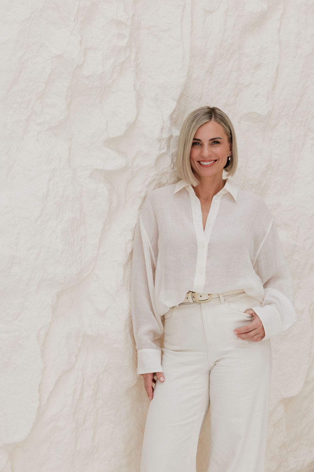 Smiling blonde woman in white linen shirt and white pants leaning against a textured white stone wall.