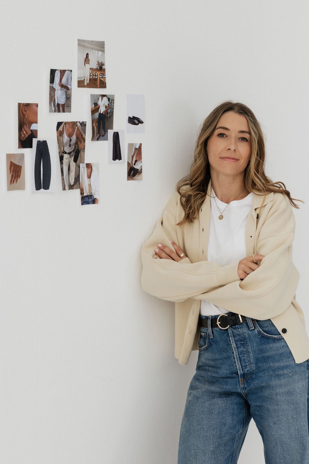 Woman in beige jacket and jeans stands arms crossed against white wall with fashion mood board photos.