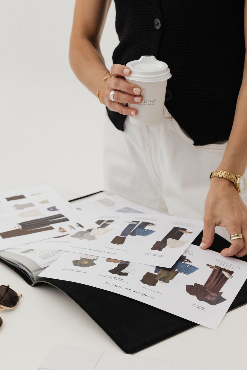 Woman in black vest holding coffee cup reviews fashion lookbook layouts spread on white table.