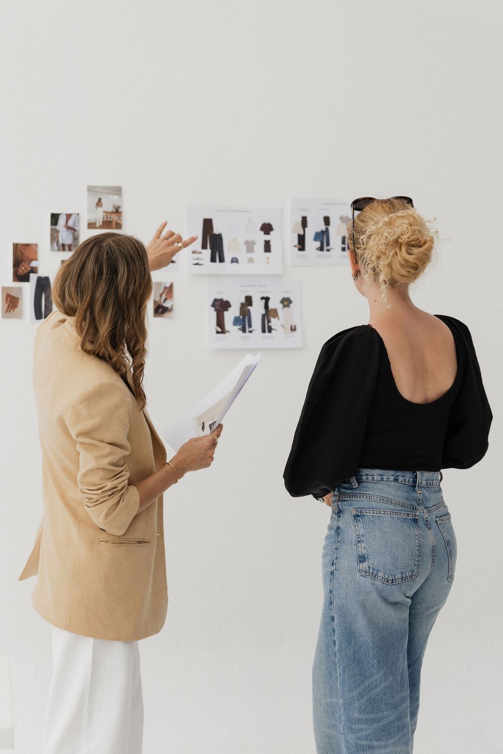 Two women reviewing fashion mood board with clothing images pinned to white wall in modern studio.