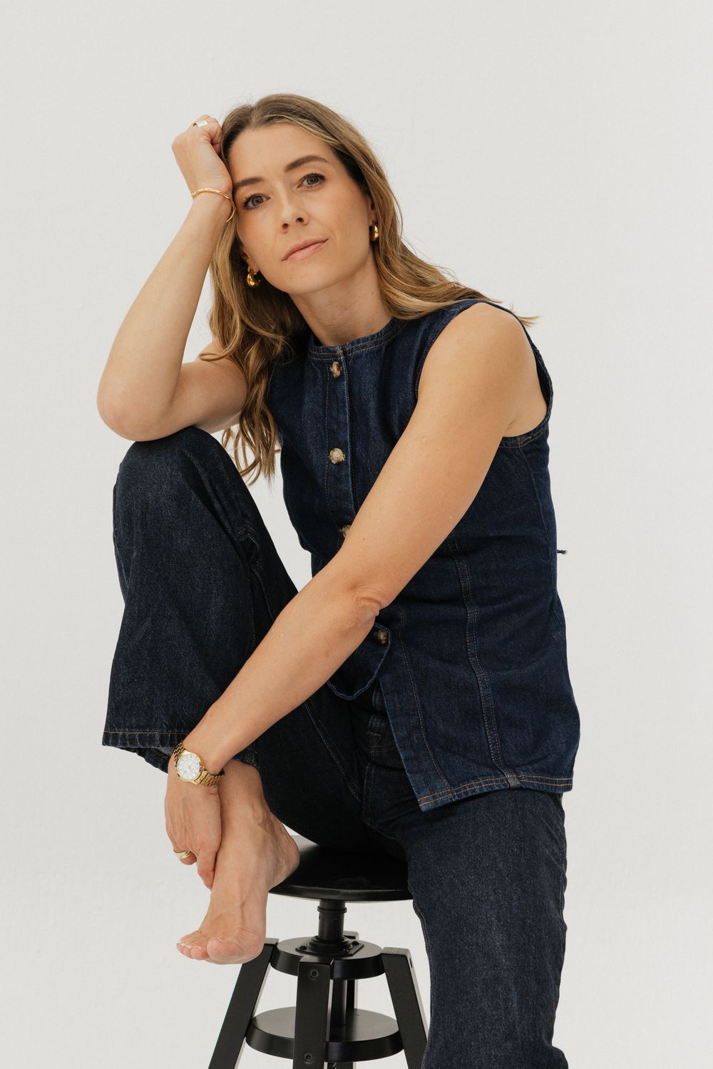Woman in dark denim vest and jeans sitting on a stool, resting her head on her hand against a white background.