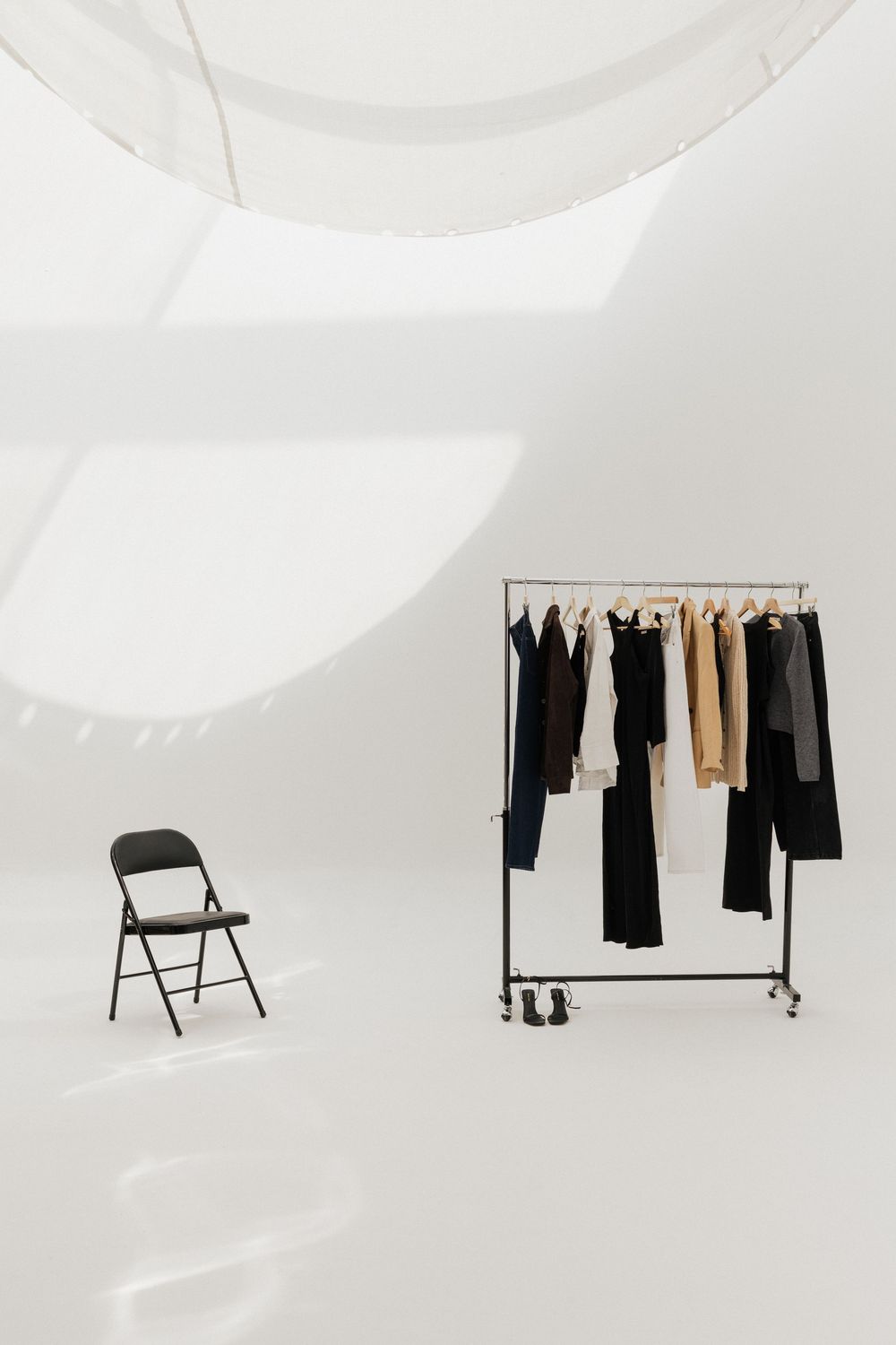 Minimalist white studio with clothing rack holding black and beige garments, beside a black folding chair.
