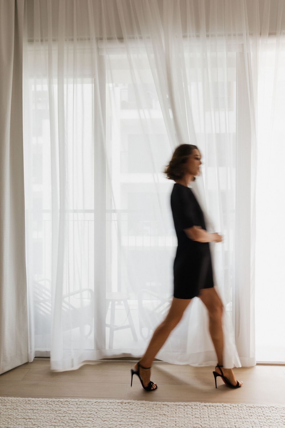 Woman in black dress and heels walking past sheer white curtains in a bright, airy room.
