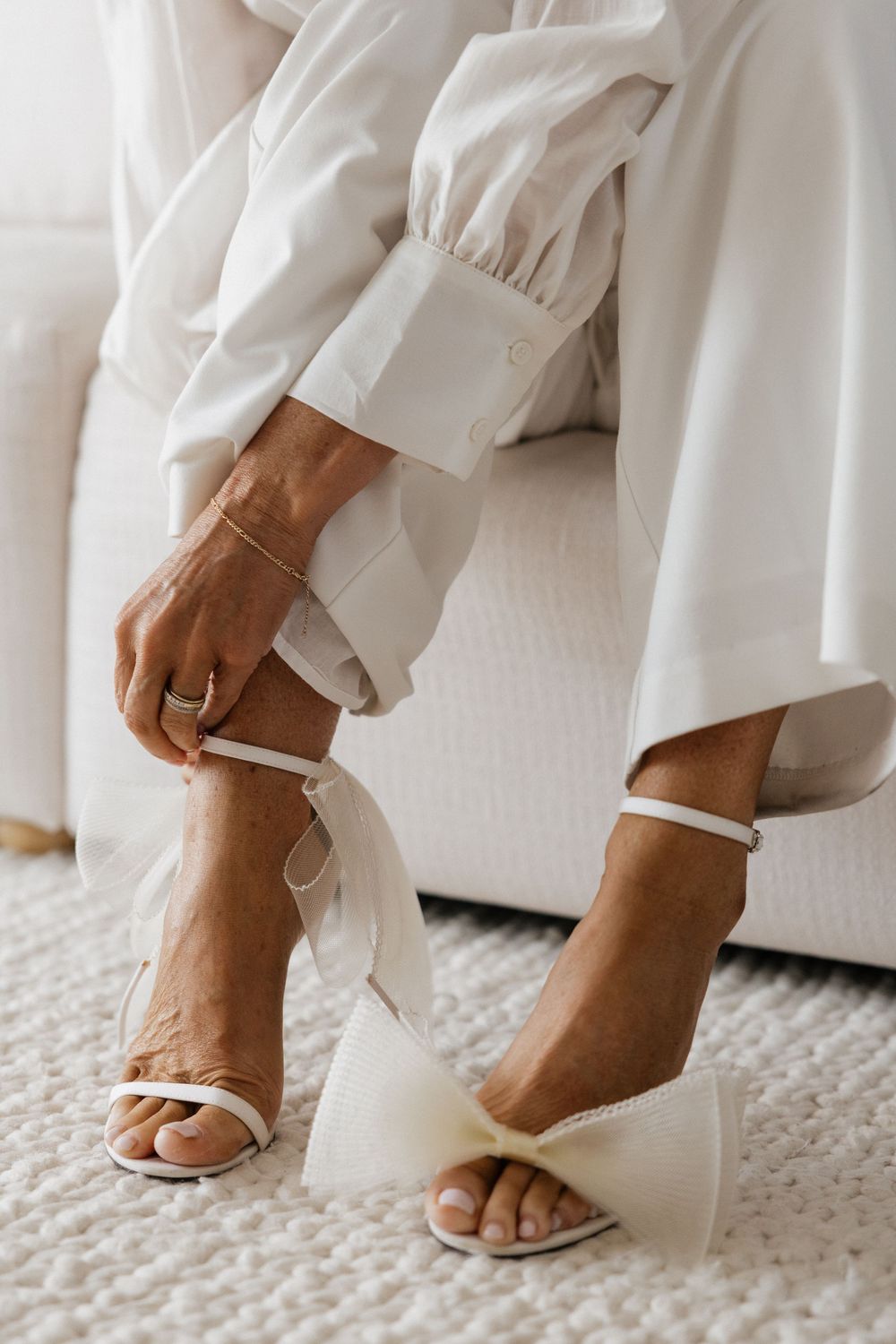 Bride putting on white heeled sandals with bow detail, wearing a white wedding dress, sitting on a white chair.