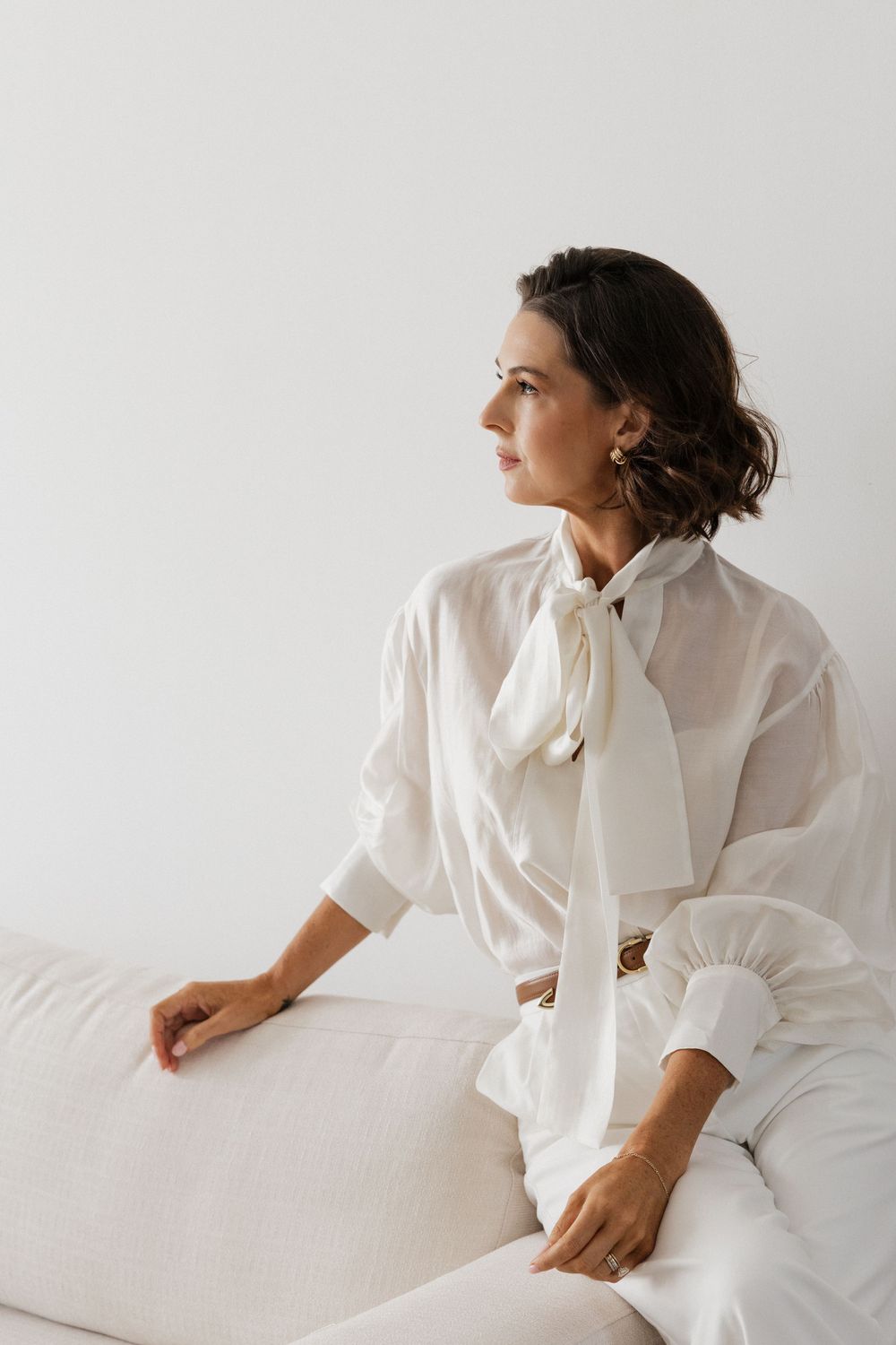 Woman in white blouse with neck tie and white pants sitting on a cream sofa against a white wall.