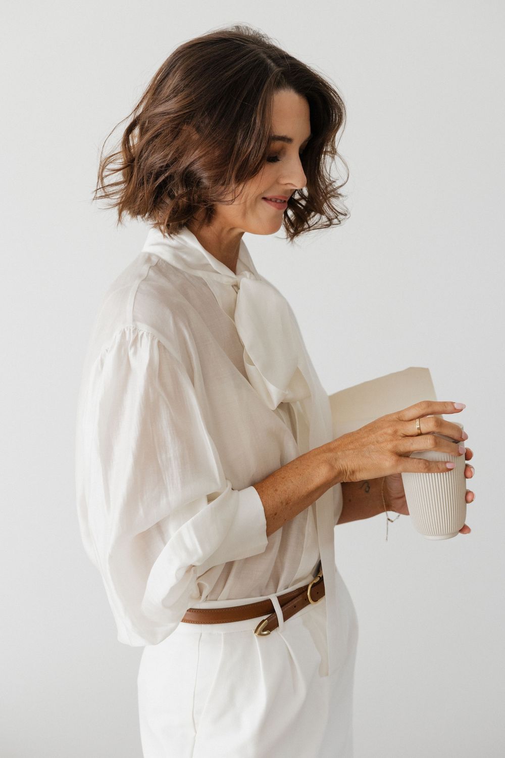 Woman in white blouse and trousers holding a coffee cup, smiling downward against a light gray background.