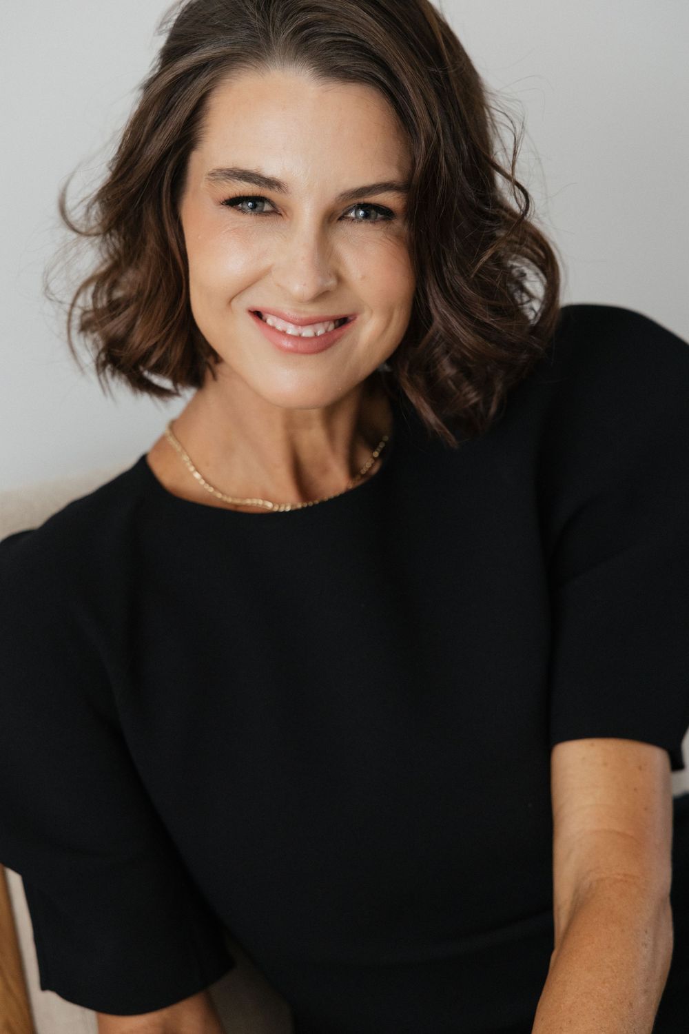 A smiling woman with short wavy brown hair wearing a black top and gold necklace poses against a light gray background.