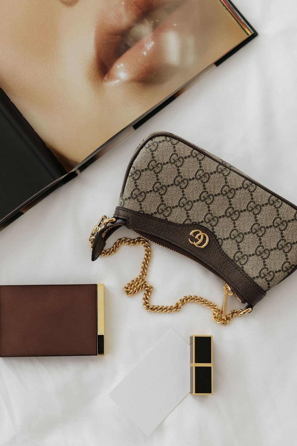 Gucci GG Supreme mini bag with gold chain strap, brown wallet, and lipstick on white surface.
