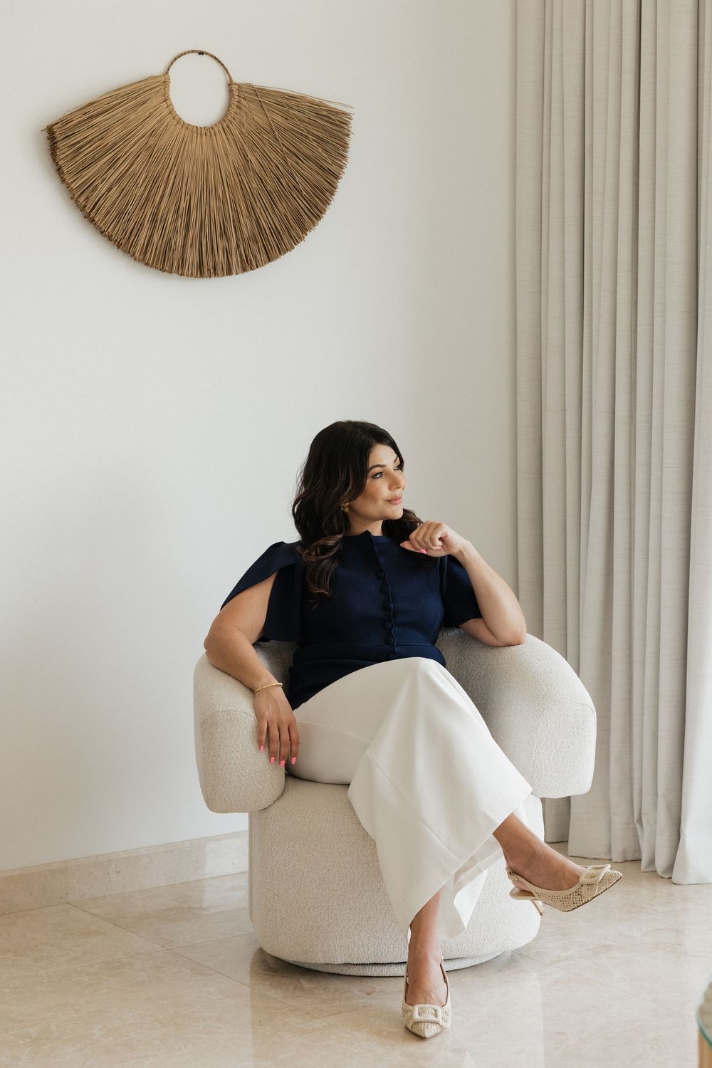Woman in navy top and white pants sits in a white chair, with a woven fan wall decor and sheer curtains behind her.