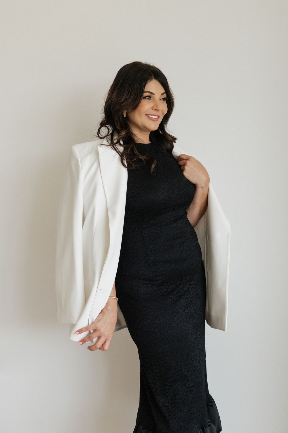 Woman in black lace gown and white blazer posing elegantly against a light background.