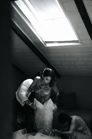 Black and white photo of bride being helped into her wedding gown in an attic room.