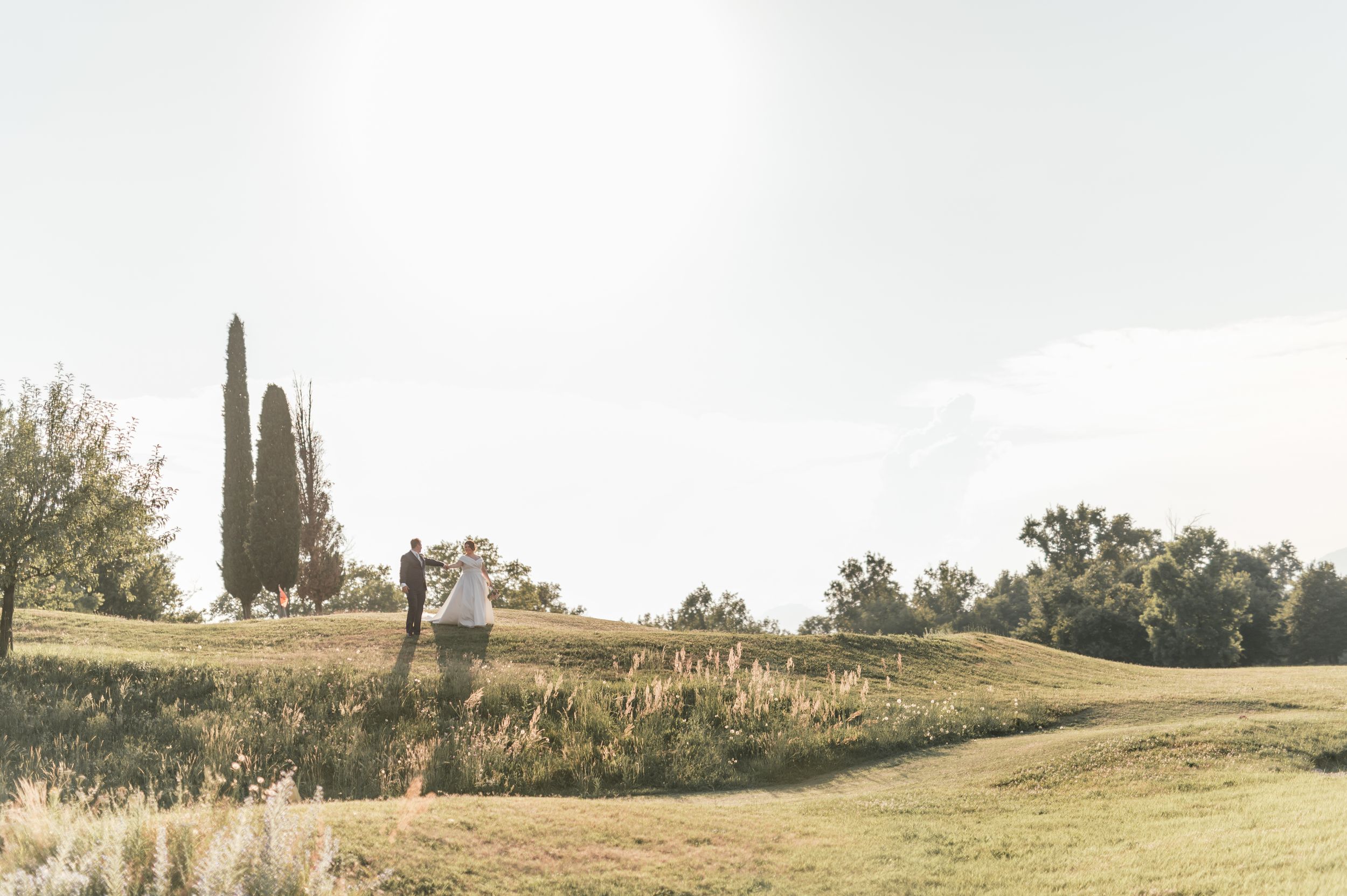 Bride and groom stroll through golden sunlit countryside landscape with tall cypress tree backdrop.