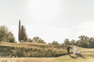 Newlyweds run hand in hand through sun-drenched Italian countryside with cypress tree in background.