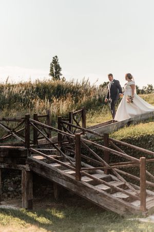Elegant bride and groom walk across rustic wooden bridge surrounded by golden meadow grasses.