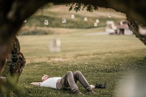 Relaxed wedding guest lies on green grass beneath tree with hanging lantern decorations above.