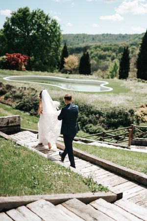 Bride and groom stroll down wooden pathway overlooking lush green landscape and pond.