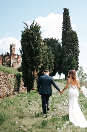 Bride and groom walk hand-in-hand toward tall cypress trees at Tuscan wedding venue.