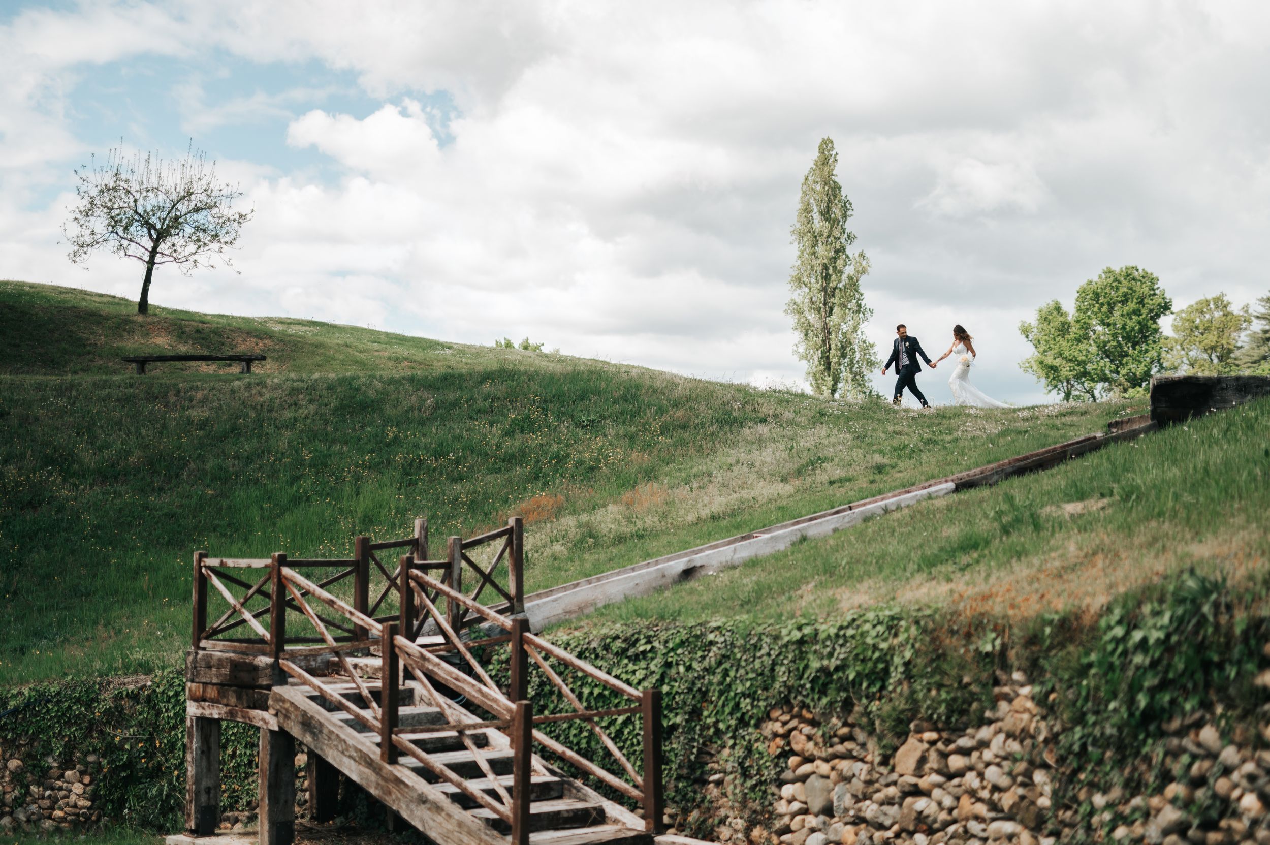 Wedding couple walks along scenic stone path over rustic wooden bridge in countryside.